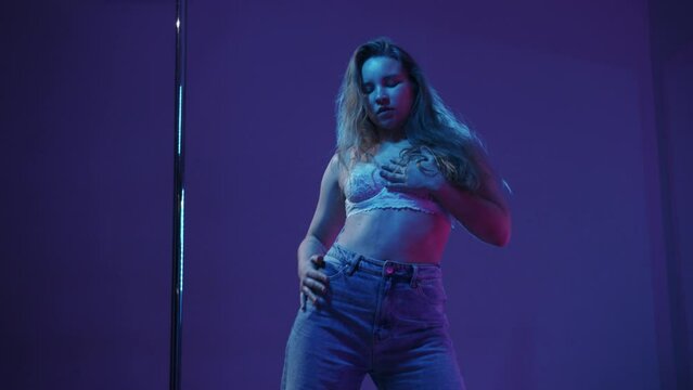 Young blonde woman in white bra dancing by the pole in blue lighting