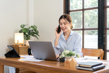 Female freelance is talking with colleague on smartphone and typing information on laptop while working