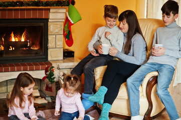 Happy young large family by a fireplace in warm living room on winter day. Mother with four kids at home read book.