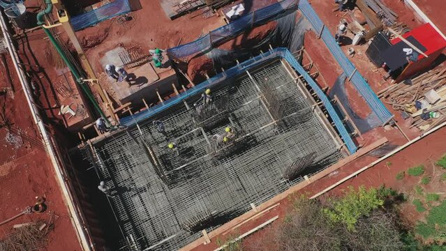 Drone sobrevoando um pr&eacute;dio sendo constru&iacute;do. Homens ao longe trabalhando na concretagem das funda&ccedil;&otilde;es, jogando cimento e espalhando. Ferros de constru&ccedil;&atilde;o instalados.