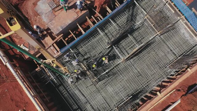 Drone sobrevoando um pr&eacute;dio sendo constru&iacute;do. Homens ao longe trabalhando na concretagem das funda&ccedil;&otilde;es, jogando cimento e espalhando. Ferros de constru&ccedil;&atilde;o instalados.