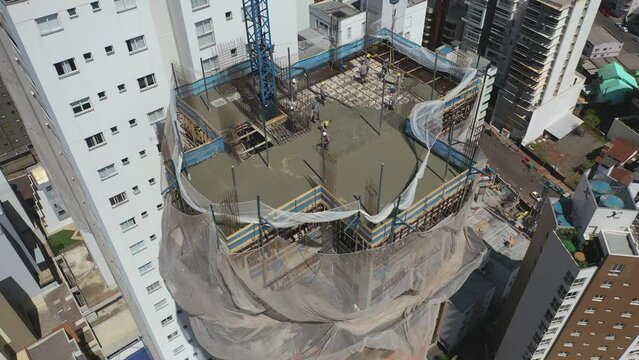 Drone sobrevoando um pr&eacute;dio sendo constru&iacute;do com uso de grua e homens ao longe trabalhando. Lage sendo executada. Cidade ao fundo.