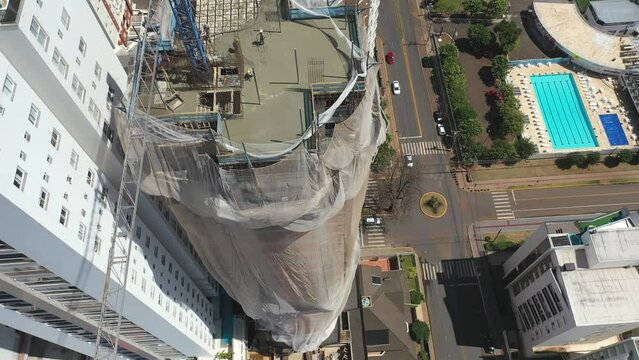 Drone sobrevoando um pr&eacute;dio sendo constru&iacute;do com uso de grua e homens ao longe trabalhando. Lage sendo executada. Cidade ao fundo.