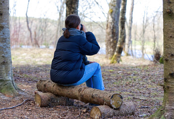 Young woman is sitting on a log bench in forest and drinking coffe. Old Wooden seat In park. close up