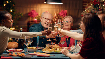 Positive festive people celebrating Christmas at evening dinner while clinking glasses with...