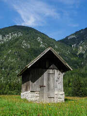 Fototapeta na wymiar Old wooden and partly stone small house in the middle of a green meadow with the Julian Alps in the background, Triglav National Park, Slovenia