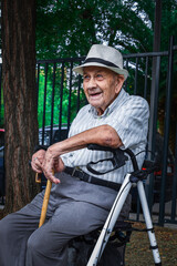 Abuelo sentado en su andador observando relajado el parque disfrutando del día con una sonrisa en la cara con su sombrero, su bastón y su andador