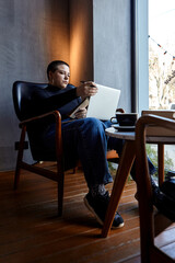 A young woman with glasses, with short hair is sitting in a cafe and working on a laptop. The girl looks at her laptop and makes notes in a notebook. The concept of distance learning