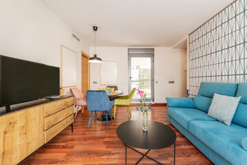 Living room of an apartment with a round wooden dining table with breakfast service with fruit and glasses with orange juice, a three-seater light blue velvet sofa,  sideboard with a flat-screen TV