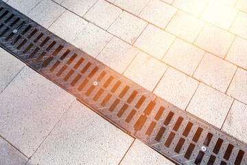 Metal storm sewer grate on sidewalk at sunset. Fragment of urban park infrastructure