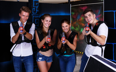 Four cheerful young male an female posing with laser pistols in their hands in dark laser tag room