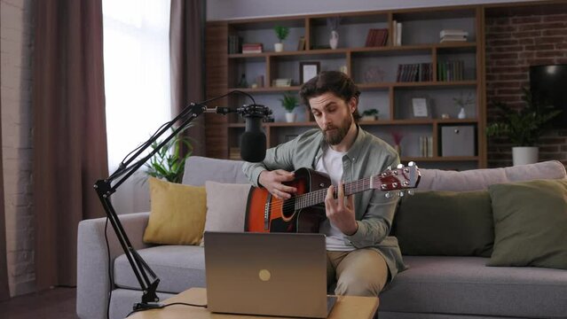 Young man recording guitar lessons for students. Online music class with distant teacher on computer. Tutor teaching music through laptop web cam. Online acoustic guitar courses, distance education.