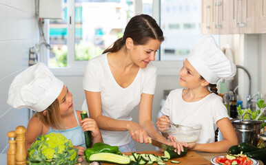 Woman with two kids in ches hats cooking together cutting and tasting vegetables for salad at home kitchen