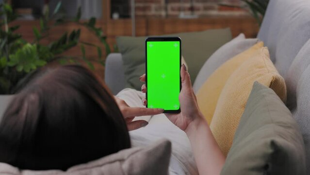 Young woman using smartphone with green mock-up screen indoors of cozy home. Scrolling through social media or online shop, swiping photos or pictures, surfing Internet,watching content videos blogs.