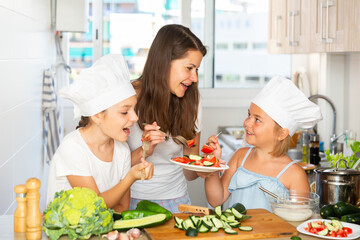Young woman with two small girls enjoying salad at kitchen after cooking