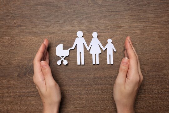 Woman protecting paper family cutout at wooden table, top view. Insurance concept