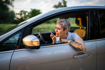 One woman mature caucasian female businesswoman sitting in car putting lipstick fixing repairing makeup on her face while waiting in summer day evening real people copy space gray hair