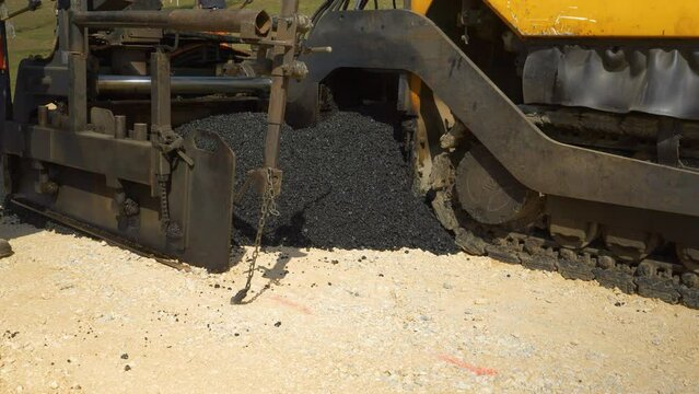 CLOSE UP: Heavy paver machinery dropping fresh black asphalt over crushed rocks. View of operating construction machinery dumping and spreading fresh blacktop surface at parking lot in morning light.