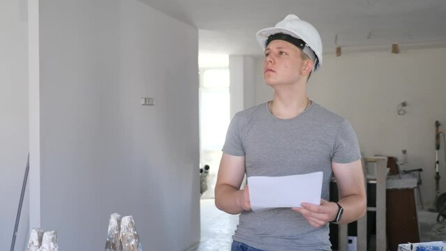 Teenage young man in hardhat doing errand in apartment during repair works, checking project documentation. Teenager learning on part time work.