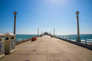 a gorgeous summer landscape at the Manhattan Beach Pier with tall lamp posts with circular lights along a blue metal hand rail with brown benches and a gorgeous clear blue sky in Manhattan Beach - Powered by Adobe