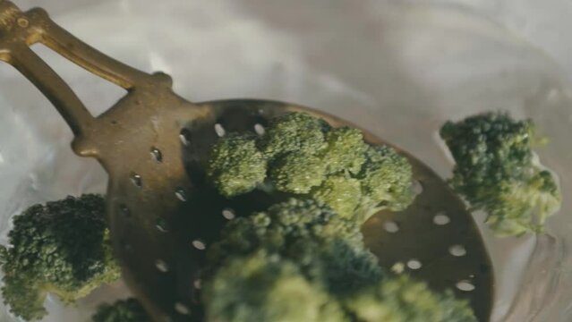 An old ladle with holes in it scoops broccoli out of the water and carries it towards the chamber, impaling it