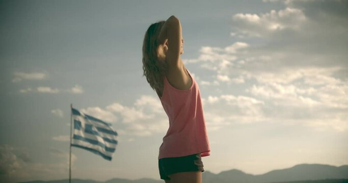 Blonde woman standing close to a Greek flag and attending around the sky touching her hair.