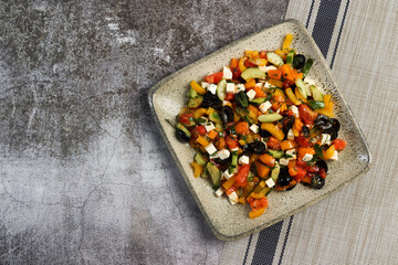 Greek salad with olives, bell peppers, tomatoes, cucumbers and feta cheese on a square  plate on a dark background. Top view, flat lay