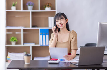 Fototapeta na wymiar Young business Asian woman working in home office, making phone call to potential client, having nice conversation, sitting at desk in front of open laptop. 