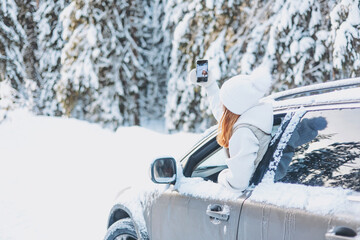 Teenager girl traveling looking out of car window and taking selfie on smartphone in winter snowy...