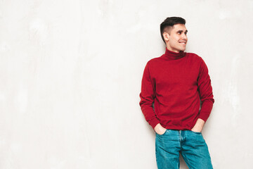 Portrait of handsome smiling model. Sexy stylish man dressed in red turtleneck sweater and jeans. Fashion hipster male posing near grey wall in studio. Isolated