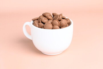 Chocolate callets in a white cup on a pastel background. Tempered chocolate confectionery concept