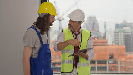 Senior engineer contractor holding tablet assigning work to young male electrician on new building,...
