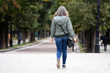 Woman in jeans and jacket walking on city street, rear view. Leisure in autumn park