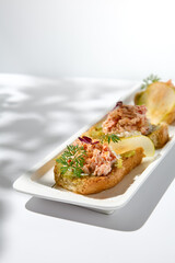 Aesthetic composition with crab bruschetta on white background with shadows from flowers. Italian bruschetta with crab, avocado and cheese on fine dining in summer. Elegant menu concept.