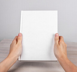 Obraz premium Hands holding closed book or diary mockup with blank white cover. Woman sitting at table and holding novel or notebook.