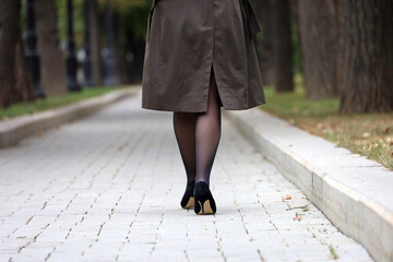 Woman in raincoat, black stockings and shoes on high heels walking in city park, rear view. Female fashion in autumn