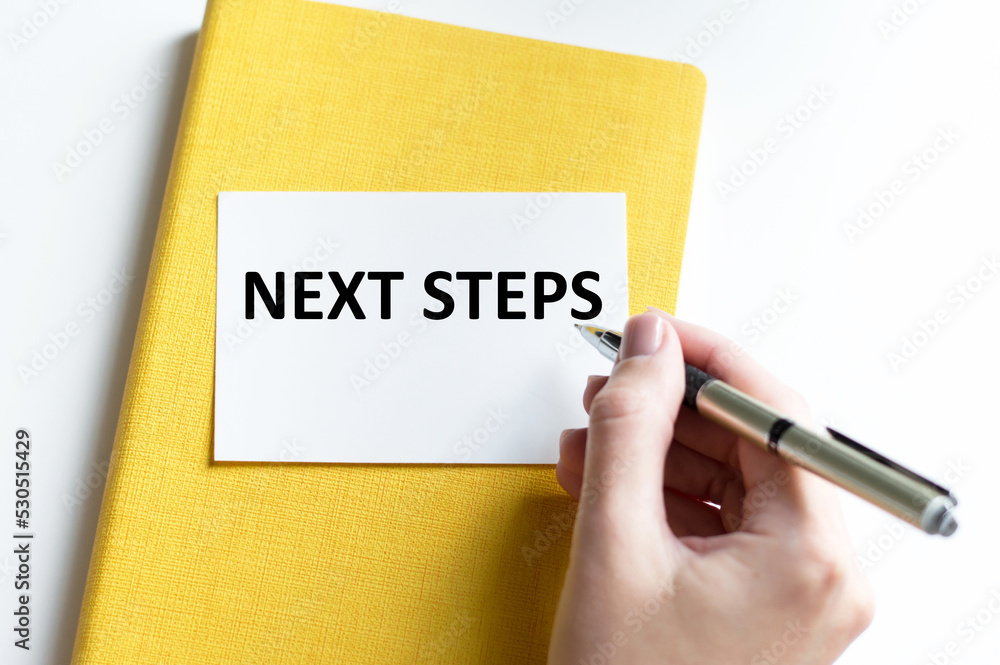 Poster next steps text on a card that lies on a notepad on a table next to a businessman's hand with a pen