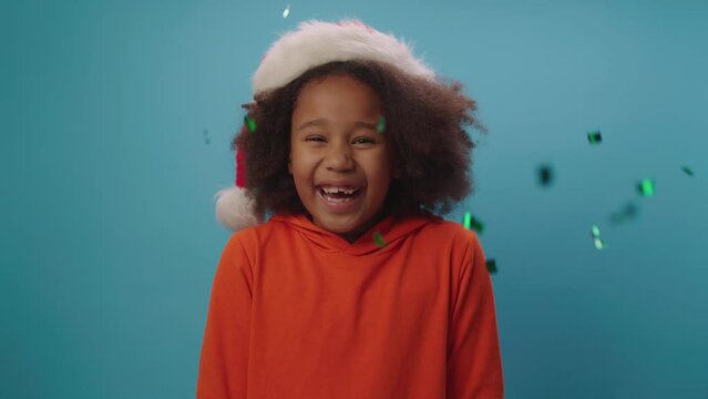 Pretty African American girl in Santa's hat with flying colour confetti. Happy school girl waiting for Christmas standing on blue background looking at camera.