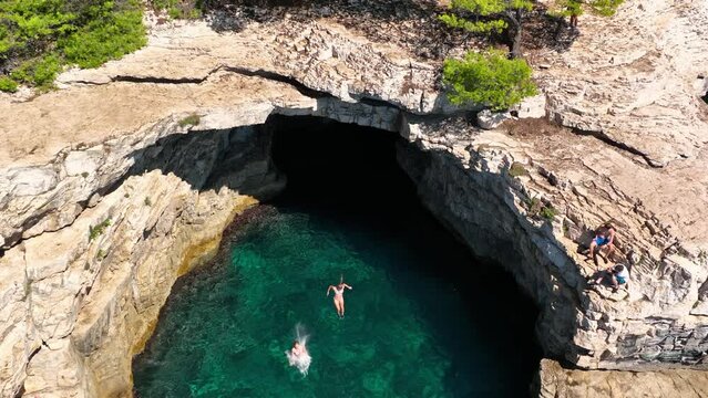 slow motion aerial view of young unrecognisable girls cliff jumping in crystal clear sea, active holiday at a paradise beach with cliffs and turquoise water, adventure vacation, cliff jumping and
