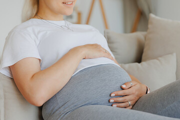 Obraz na płótnie Canvas Pregnant woman in pajamas holds belly while sitting on gray sofa. Scandinavian home pregnancy concept