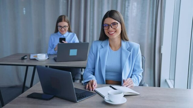 Happy female office employees working in a light room. Women sit at desks using laptops and smartphones. Blurred backdrop.