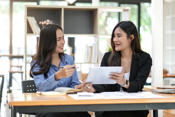 Two pretty young Asian businesswoman sitting at desk with laptop doing paperwork together discussing project financial report. Corporate business collaboration concept.