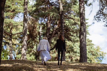 Fototapeta premium Two women together - blonde in light and redhead in dark clothes - walking in a forest, back view. Cosplay - female version of Angel and Demon (Good Omens)