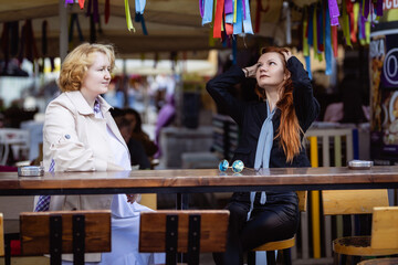 Two women together - blonde in light and redhead in dark clothes talking at the bar of a street cafe. Photo shoot based on the series 