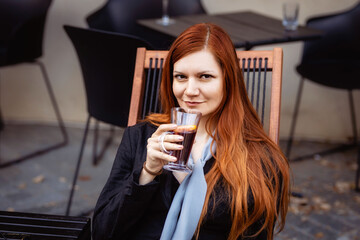 Girl drinking a hot drink while sitting in a street cafe. Portrait of a slender red-haired woman in a business black suit.