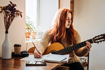 Unaltered candid portrait of young red haired woman in sweater playing acoustic guitar sitting by window at home. Hobbies, indoor home activities for adults in winter, autumn. - obrazy, fototapety, plakaty