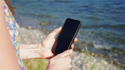 Close-up Mockup image of a woman's hands holding black phone with blank desktop screen while taking a photo by the sea.