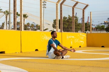 Young African American male in a blue shirt and overalls meditating on a yellow court