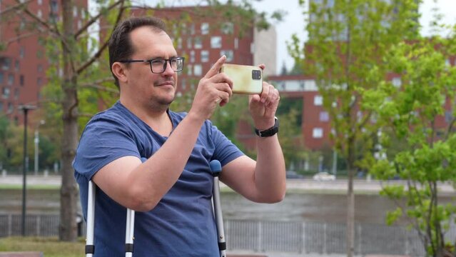 Mature plus size man with crutches and glasses exploring city on vacation. Handicapped man taking picture of park on  phone while sightseeing in town.
