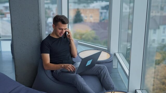 Young male freelancer using computer and phone at once. Busy self-employed man working in a comfortable place at the window. High angle view.